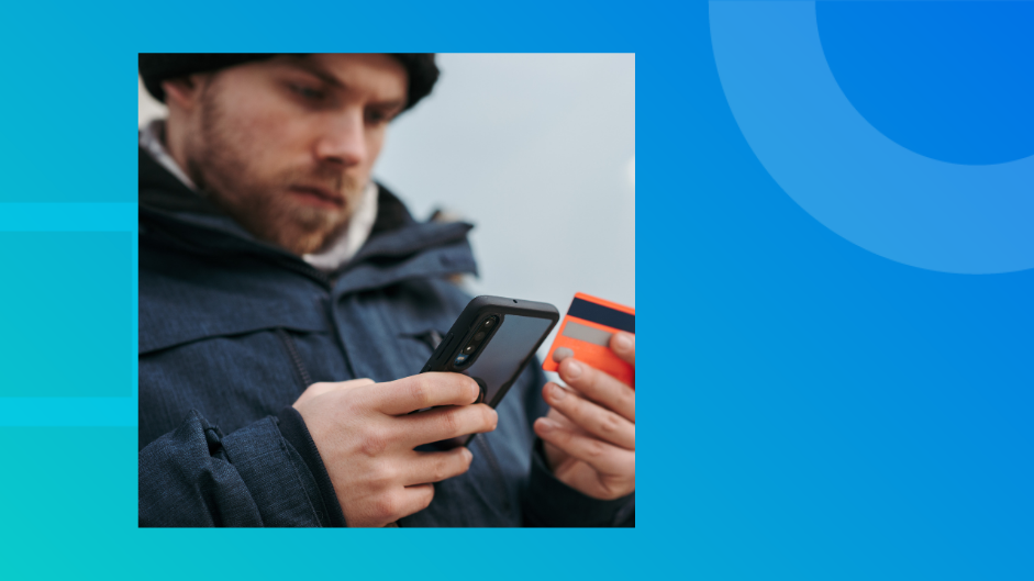A man in a winter jacket checks his phone while holding a credit card outdoors, representing US consumers managing rising credit card debt in 2026